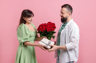 Kırmızı gül buketini ve hediye kutusunu pembe arka plandaki sevgilisine sunan bir adam.