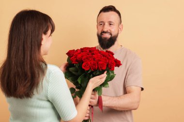 Adam bej arka planda sevgilisine kırmızı gül buketi sunuyor.