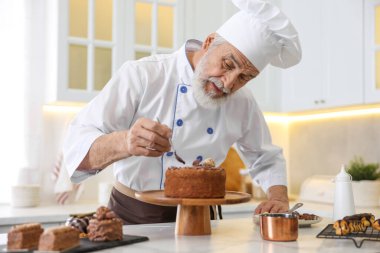 Bir kepçe erimiş çikolatayla yaşlı pasta şefi mutfaktaki masada pasta yapıyor.