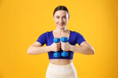 Spor kıyafetli bir kadın turuncu arka planda dambıllarla egzersiz yapıyor.