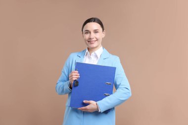 Emlakçının elinde ev şeklindeki anahtarlık ve bej arka planda klasör var.