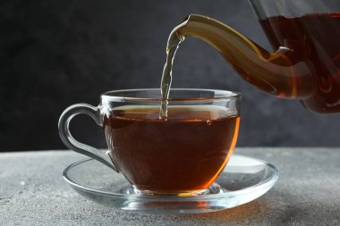 Gri masada çaydanlıktan fincana taze demlenmiş çay dökülüyor, yakın plan.