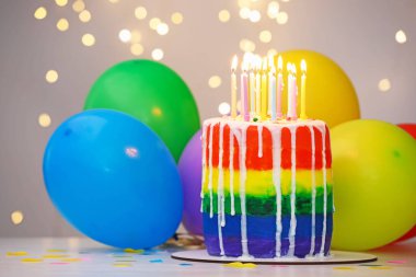 Lezzetli gökkuşağı doğum günü pastası, yanan mumlar, renkli balonlar ve beyaz masanın üzerindeki konfetiler, gri arka planda bulanık ışıklar, yakın plan