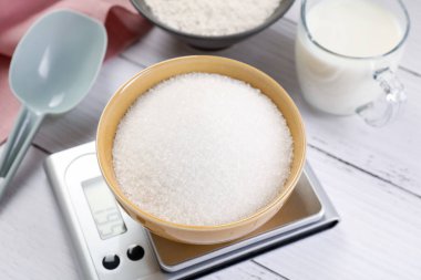 Dijital mutfak terazisi, bir kase şeker ve beyaz ahşap masa üzerindeki diğer ürünler, yakın plan.