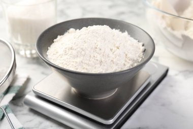 Dijital mutfak terazisi, beyaz mermer masa üzerinde bir kase un ve diğer ürünler, yakın plan.