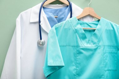 Açık yeşil arka planda askılara steteskop takılmış sağlık görevlileri üniformaları, yakın plan.