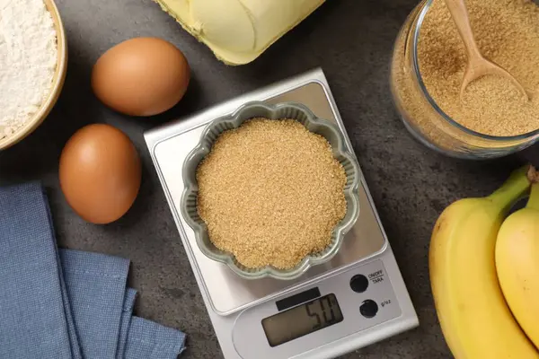 Mutfak terazisinde bir kase un ve diğer ürünler gri masada, düz yerde.