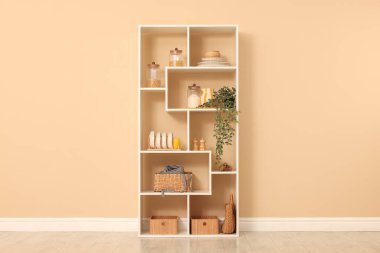 Shelving unit with dishware, jars of different products and other stuff near beige wall indoors