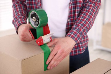 Karton kutuyu kapalı bir yere kapatmak için yapıştırıcılı bant kullanan bir adam.