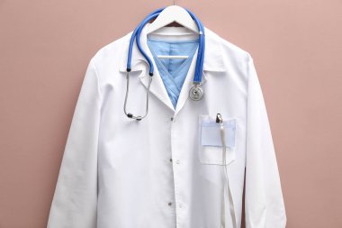 Doktor ceketi, steteskop ve rozeti bej arka planda asılı.