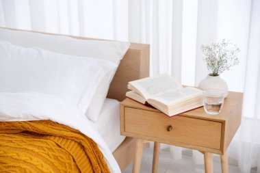 İçinde kitap olan komodin, vazoda çiçekler, yatağın yanında bir bardak su ve bardaklar.