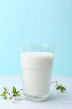 Açık mavi arka planda beyaz fayanslı masada bir bardak taze süt ve çiçek.