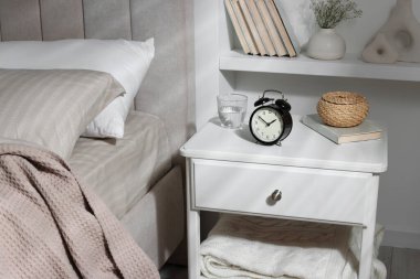 Alarm saati, bir bardak su, hasır kutu ve yatağın yanında kitap olan komodin.