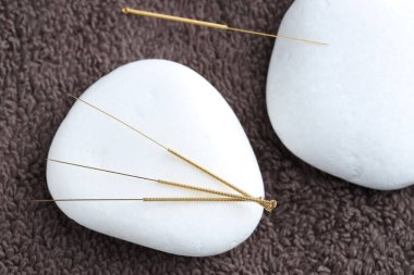 Akupunktur iğneleri ve spa taşları kahverengi havluda, düz yerde.