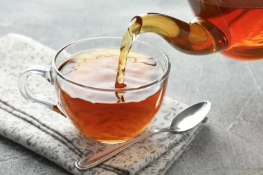 Gri masada çaydanlıktan fincana taze demlenmiş çay dökülüyor, yakın plan.