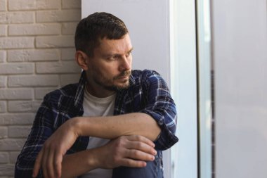 Evdeki pencerenin yanında oturan depresif bir adam.