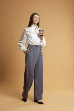 Şık giyinmiş, bej arka planda bir fincan kahveyle güzel bir kadın.