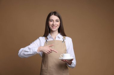 Bej arka planda bir fincan sıcak içeceğe işaret eden gülümseyen baristanın portresi