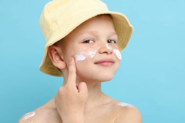 Küçük çocuk açık mavi arka planda cildine güneş kremi sürüyor.