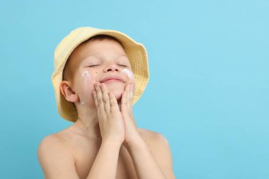 Küçük çocuk açık mavi arka planda cildine güneş kremi sürüyor.