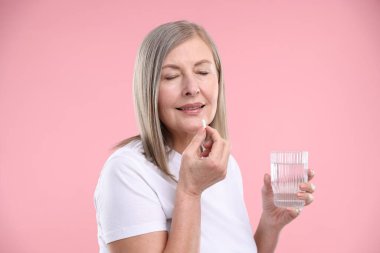Bir bardak suyla mutlu bir kadın pembe arka planda kolajen hapı alıyor.