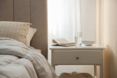 Yatağın yanında kitap, kase ve bir bardak su olan komodinin yanında.