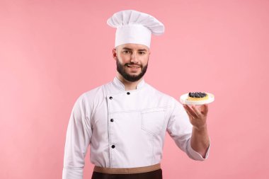 Üniformalı mutlu pastacı pembe arka planda yaban mersinli tart tutuyor.