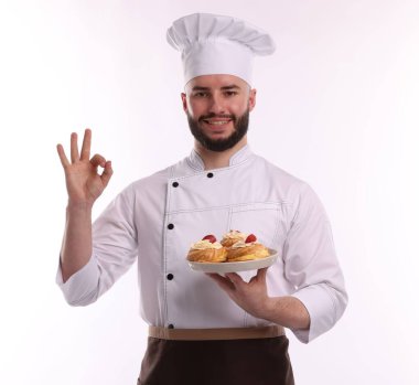 Üniformalı mutlu pastacı elinde çilekli lezzetli profiteroller tutuyor ve beyaz arka planda güzel bir jest yapıyor.