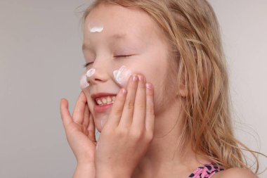 Gülümseyen küçük kız, gri arka planda yüzüne güneş kremi sürüyor.