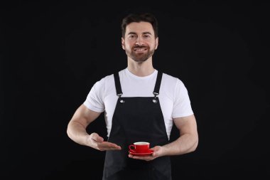 Siyah arka planda bir fincan sıcak içecekle gülümseyen baristanın portresi.