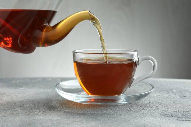 Gri masada çaydanlıktan fincana taze demlenmiş çay dökülüyor, yakın plan.