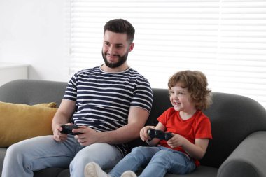 Mutlu baba ve oğlu evdeki kanepede oyun kumandalarıyla oynuyorlar.