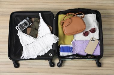 Farklı kıyafetler, akıllı telefon, kamera ve güneş gözlükleri açık bavulda ahşap zemin, üst manzara