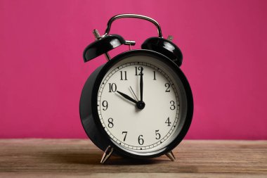 Ahşap masada parlak pembe arka planda siyah bir çalar saat.