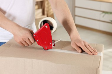 Karton kutuyu kapalı bir yere kapatmak için yapıştırıcılı bant kullanan bir adam.
