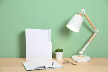 Lambalı, kırtasiyeli ve açık yeşil duvarlı ahşap masadaki bitkili şık bir iş yeri.