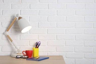 Farklı kırtasiye malzemeleri, kahve, lamba ve bardaklar beyaz tuğla duvarın yanındaki ahşap ofis masasında.