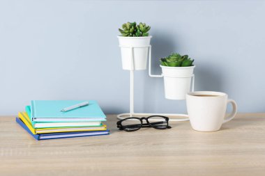 Farklı kırtasiye malzemeleri, bardaklar, kahve ve çiçeklerle aydınlık duvarın yanındaki ahşap ofis masasında.