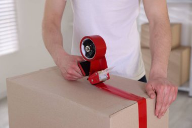 Karton kutuyu kapalı bir yere kapatmak için yapıştırıcılı bant kullanan bir adam.