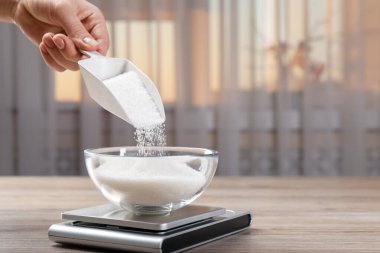 İçerideki ahşap masada kaseye şeker ve dijital mutfak terazisi ekleyen kadın, yakın plan. Metin için boşluk