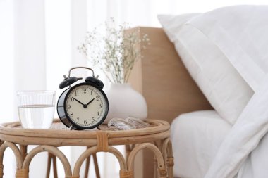 Komodinin yanında çalar saat, vazoda çiçekler, yatağın yanında bir bardak su ve bardaklar, yakın plan.