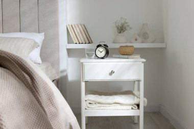 Alarm saati, bir bardak su, hasır kutu ve yatağın yanında kitap olan komodin.