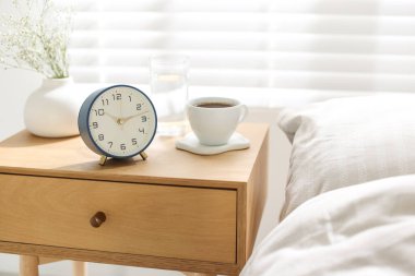 Çalar saati olan bir komodin, vazoda çiçekler, yatağın yanında bir bardak su ve kahve, yakın plan.