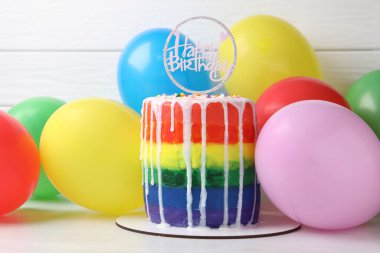 Lezzetli gökkuşağı doğum günü pastası. Masada beyaz ahşap arka planda balonlar var.