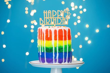 Lezzetli gökkuşağı doğum günü pastası, üstünde tepesi olan açık mavi arka plan, bulanık ışıklar, yakın plan.