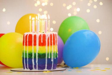 Lezzetli gökkuşağı doğum günü pastası, yanan mumlar, renkli balonlar ve beyaz masanın üzerindeki konfetiler, gri arka planda bulanık ışıklar, yakın plan