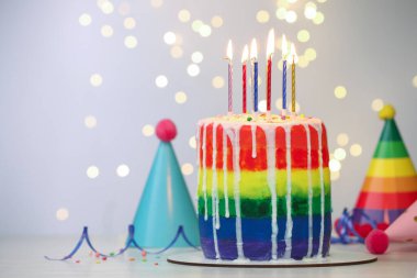 Enfes gökkuşağı doğum günü pastası, yanan mumlar, flamalar, serpiştirmeler ve parti şapkaları beyaz masanın üzerinde bulanık ışıklı arka planda, yakın plan. Metin için boşluk