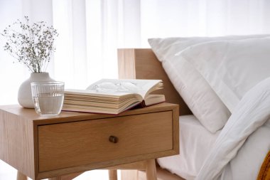 İçinde kitap olan komodin, vazoda çiçekler, yatağın yanında bir bardak su ve bardaklar.