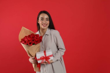 Kırmızı arka planda lale buketi ve hediye kutusu olan mutlu bir kadın. Mesaj için yer var. 8 Mart kutlamaları