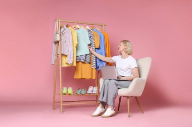 Gülümseyen moda tasarımcısı pembe arka planda dizüstü bilgisayarı ve elbise askısı var.